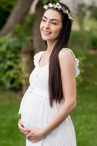 Belle femme enceinte heureuse en robe blanche et couronne florale toucher le ventre et regarder loin dans le parc — Photo de stock