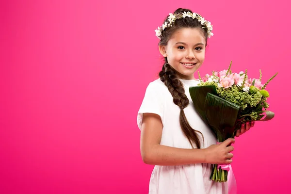 Heureux enfant en robe blanche et couronne florale tenant bouquet de fleurs et souriant à la caméra isolé sur rose — Photo de stock