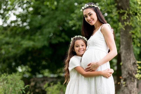 Enfant heureux embrassant mère enceinte et souriant à la caméra dans le parc — Photo de stock