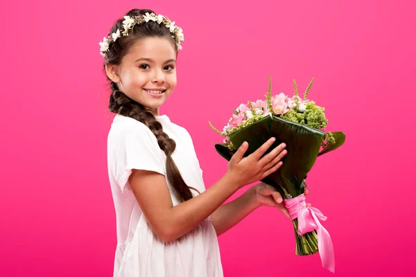 Adorable child in white dress and floral wreath holding flower bouquet and smiling at camera isolated on pink — Stock Photo