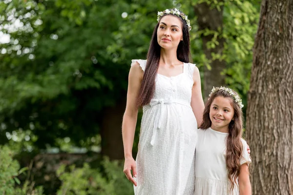 Happy pregnant mother and cute daughter in white dresses and wreaths standing together and looking away in park — Stock Photo