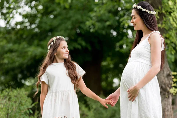 Madre embarazada feliz y linda hija en vestidos blancos y guirnaldas tomados de la mano y sonriendo entre sí en el parque - foto de stock