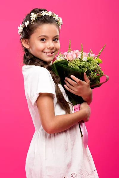 Adorabile bambino felice in abito bianco e corona floreale tenendo bouquet di fiori e sorridendo alla fotocamera isolata su rosa — Foto stock