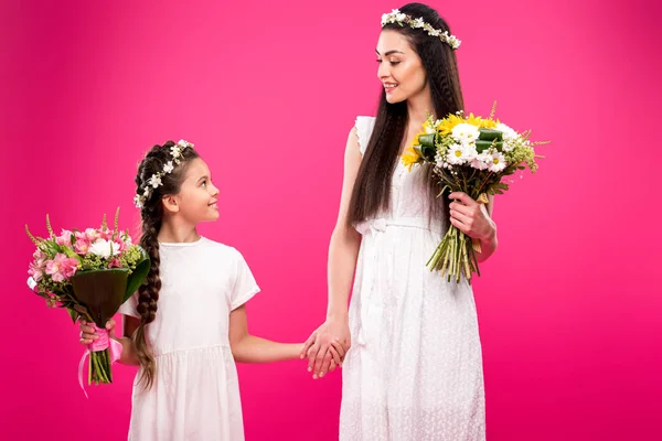 Hermosa madre feliz e hija en vestidos blancos y guirnaldas florales sosteniendo ramos y sonriendo unos a otros aislados en rosa - foto de stock