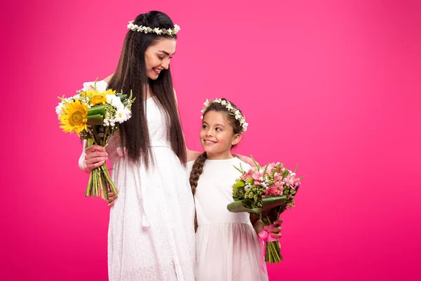 Beautiful happy mother and daughter in white dresses and floral wreaths holding bouquets isolated on pink — Stock Photo