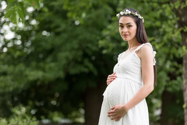 Beautiful pregnant woman in white dress and floral wreath touching belly and smiling at camera in park — Stock Photo