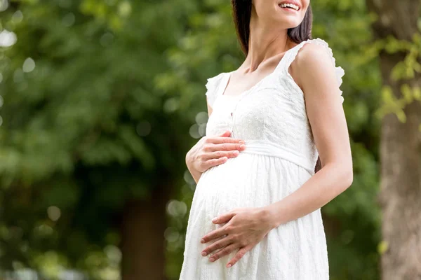 Cortado tiro de mulher grávida sorridente em vestido branco tocando barriga no parque — Fotografia de Stock