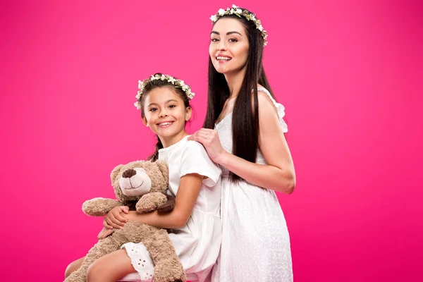 Beautiful happy mother and adorable daughter with teddy bear smiling at camera isolated on pink — Stock Photo