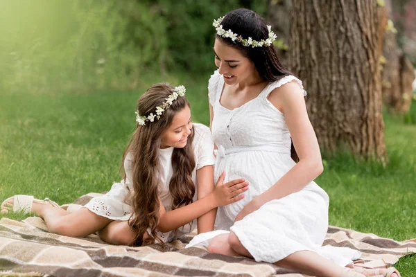 Милая улыбающаяся дочь трогает живот беременной матери, сидя вместе на клетке в парке — стоковое фото