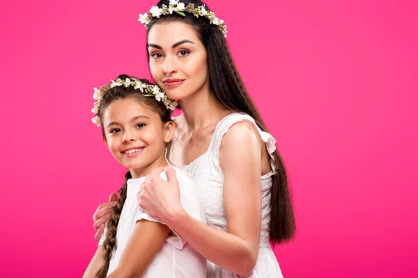 Beautiful happy mother and daughter in white dresses and floral wreaths smiling at camera isolated on pink — Stock Photo