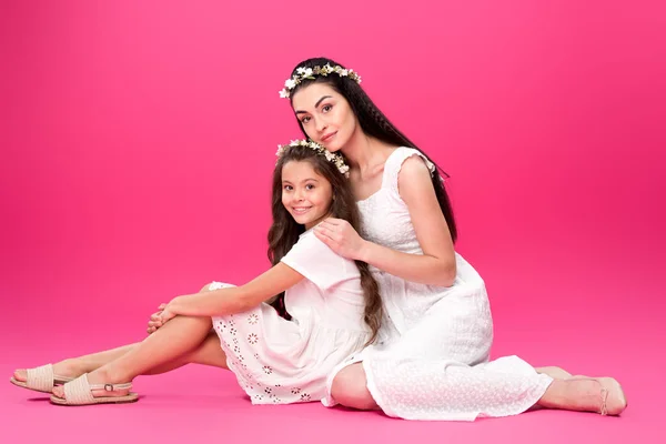 Vue pleine longueur de belle mère heureuse et fille en robes blanches et des couronnes florales assis ensemble et souriant à la caméra sur rose — Photo de stock