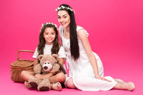 Hermosa madre feliz e hija en vestidos blancos sentados con oso de peluche y cesta de picnic en rosa - foto de stock