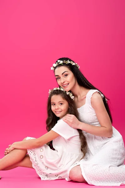 Hermosa feliz madre e hija en vestidos blancos sentados juntos y sonriendo a la cámara en rosa - foto de stock
