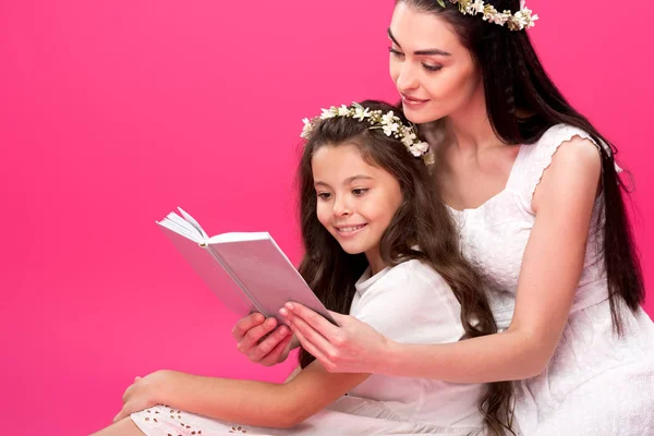 Beautiful mother and daughter in white dresses sitting together and reading book isolated on pink — Stock Photo