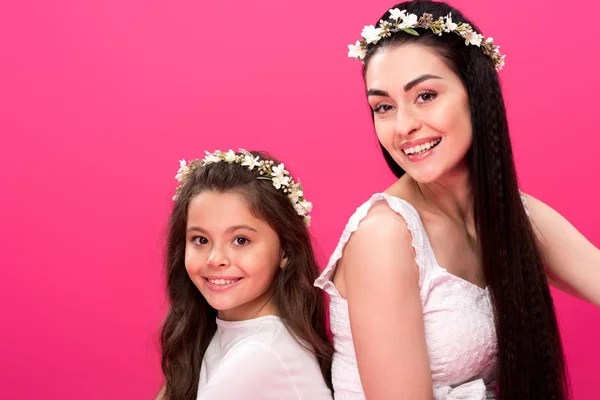Hermosa madre feliz e hija en vestidos blancos y guirnaldas florales sonriendo a la cámara aislada en rosa - foto de stock
