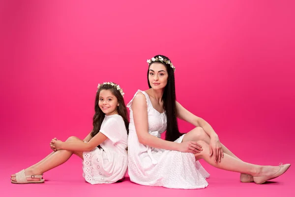 Beautiful mother and daughter in white dresses sitting back to back and smiling at camera on pink — Stock Photo