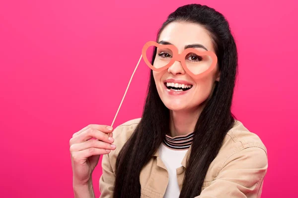 Glückliche junge Frau mit herzförmiger Brille auf Party-Stick und lächelnd in die Kamera vereinzelt auf rosa — Stockfoto