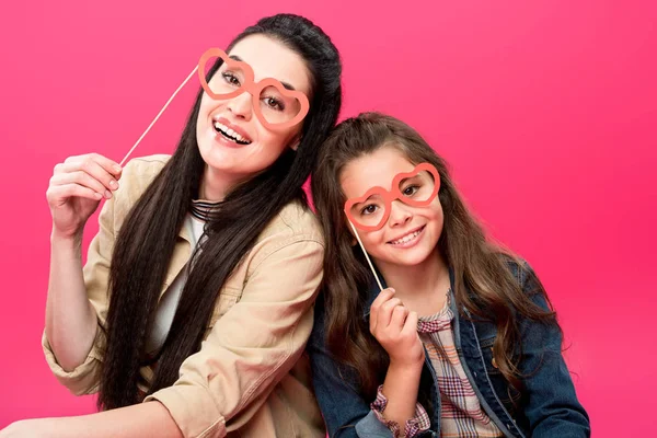 Happy mother and daughter holding heart shaped eyeglasses on sticks and smiling at camera isolated on pink — Stock Photo