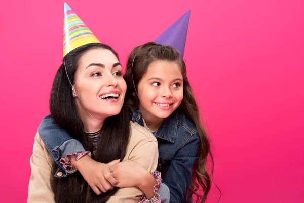 Happy mother and daughter in party hats hugging and looking away isolated on pink — Stock Photo