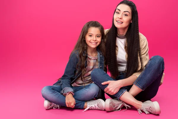 Visão de comprimento total de bela mãe feliz e filha sentados juntos e sorrindo para a câmera em rosa — Fotografia de Stock