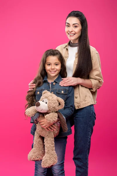 Madre y linda hijita con oso de peluche de pie juntos y sonriendo a la cámara aislada en rosa - foto de stock