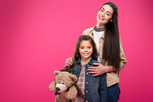 Feliz madre y linda hijita con osito de peluche de pie juntos y sonriendo a la cámara aislado en rosa - foto de stock
