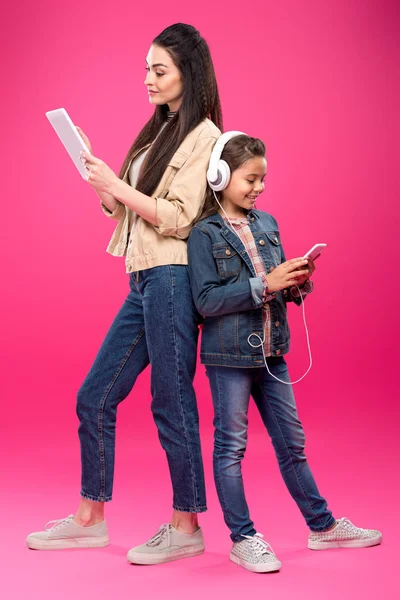Vista lateral de la madre y la hija sonrientes en auriculares de pie espalda con espalda y el uso de dispositivos digitales en rosa - foto de stock