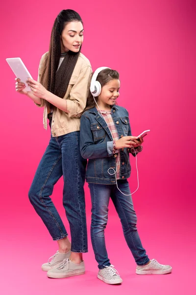 Vista completa de la madre sonriente sosteniendo la tableta digital y mirando a la hija en los auriculares usando el teléfono inteligente en rosa - foto de stock