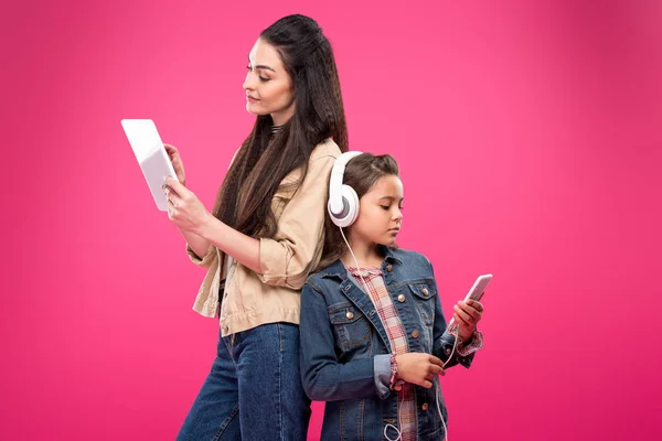 Mãe e filha em fones de ouvido de pé para trás e usando dispositivos digitais isolados em rosa — Fotografia de Stock