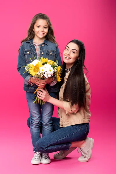 Full length view of happy young mother and adorable daughter with flower bouquet smiling at camera on pink — Stock Photo