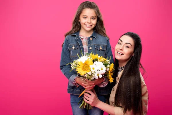 Hermosa madre feliz y la hija sosteniendo ramo de flores y sonriendo a la cámara aislada en rosa - foto de stock