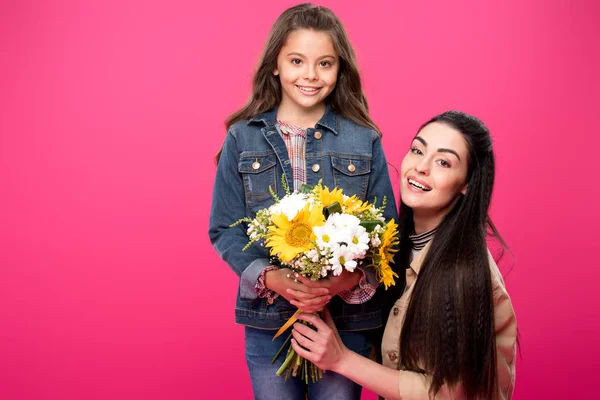 Bela mãe feliz e filha segurando buquê de flores e sorrindo para a câmera isolada em rosa — Fotografia de Stock