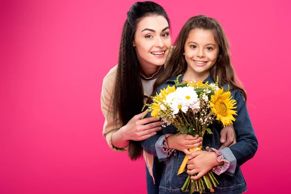 Mãe feliz e filha segurando belo buquê de flores e sorrindo para a câmera isolada em rosa — Fotografia de Stock