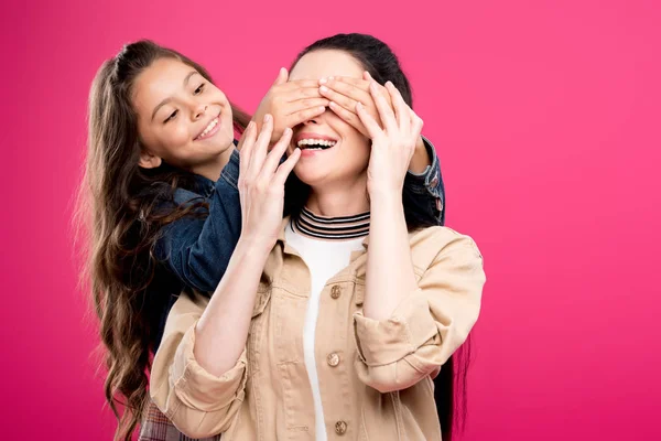 Sonriente hija cerrando los ojos a feliz madre aislado en rosa - foto de stock