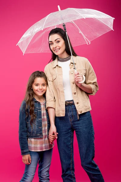 Madre con paraguas y adorable hija feliz cogida de la mano y sonriendo a la cámara aislada en rosa - foto de stock