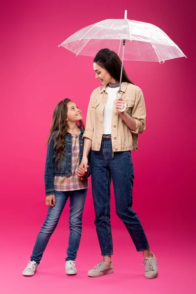Feliz hija y madre con paraguas tomados de la mano y sonriéndose mutuamente en rosa - foto de stock