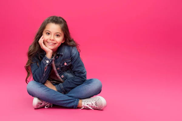 Чарівна щаслива дитина в джинсовому одязі, сидячи з рукою на підборідді і посміхаючись на камеру на рожевому — стокове фото