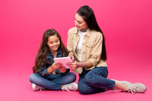 Hermosa madre feliz e hija sentados juntos y usando tableta digital en rosa - foto de stock
