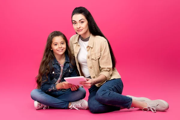 Schöne glückliche Mutter und Tochter sitzen zusammen mit dem digitalen Tablet und lächeln in die Kamera auf rosa — Stockfoto
