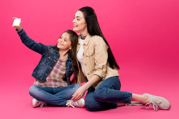 Happy mother and daughter sitting together and taking selfie with smartphone on pink — Stock Photo