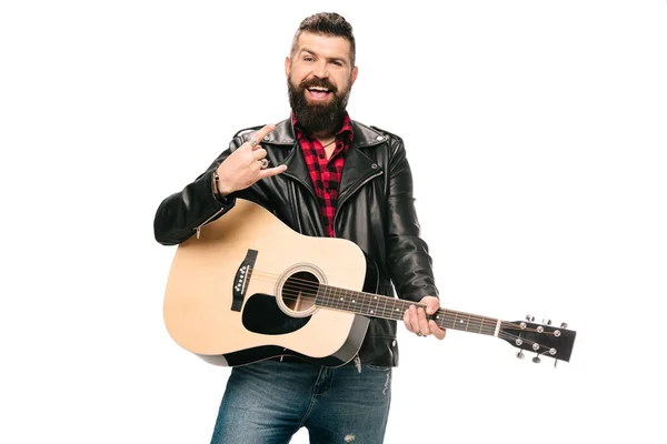 Handsome smiling musician in black leather jacket holding acoustic guitar and showing rock and roll sign, isolated on white — Stock Photo