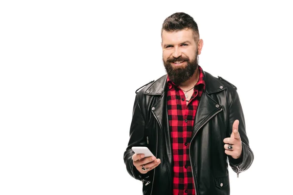 Glücklicher bärtiger Mann in schwarzer Lederjacke mit Zeigefinger und Smartphone, isoliert auf weiß — Stockfoto