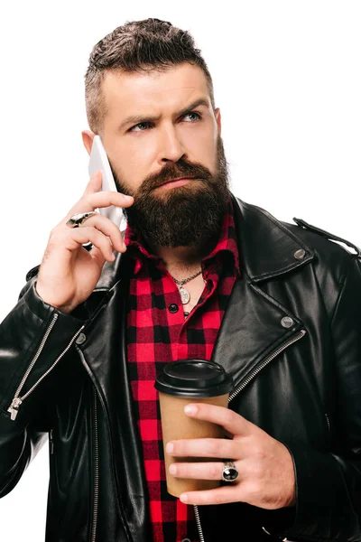 Hombre barbudo guapo en chaqueta de cuero negro sosteniendo café para ir y hablar en el teléfono inteligente, aislado en blanco - foto de stock