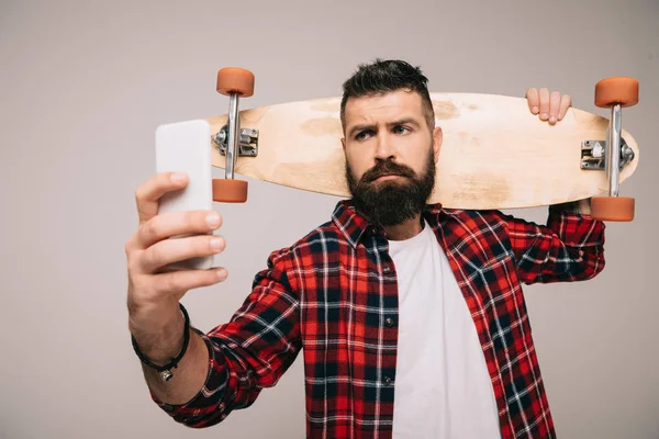 Hombre barbudo guapo con camisa a cuadros sosteniendo longboard y tomando selfie en el teléfono inteligente aislado en gris - foto de stock