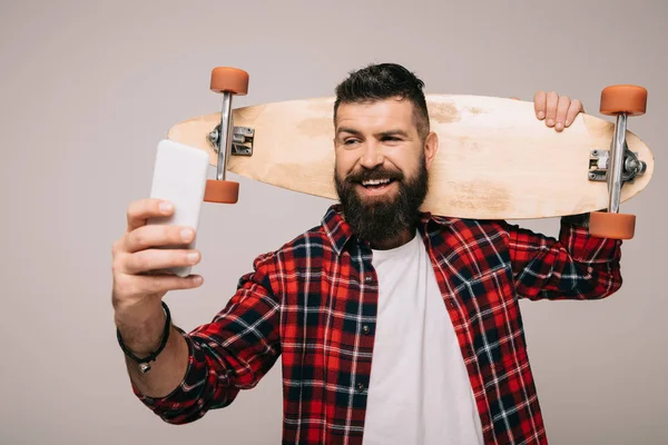 Uomo allegro in camicia a scacchi tenendo longboard e scattando selfie su smartphone isolato su grigio — Foto stock