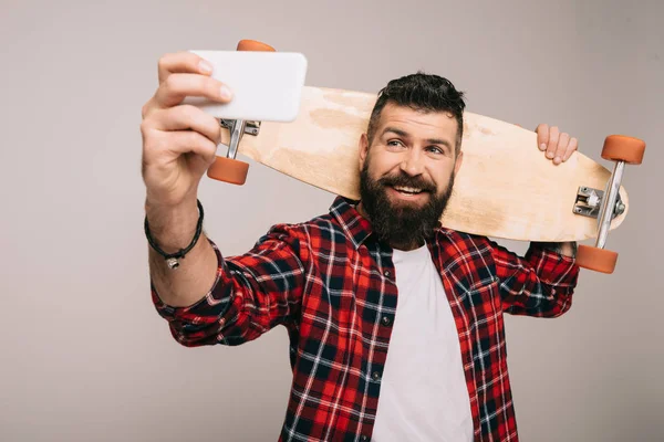 Улыбающийся бородатый мужчина в клетчатой рубашке, держащий скейтборд и делающий селфи на смартфоне, изолированном на сером — стоковое фото