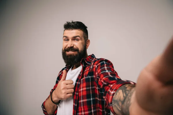 Sonriente hombre barbudo con camisa a cuadros mostrando el pulgar hacia arriba mientras toma selfie, aislado en gris - foto de stock