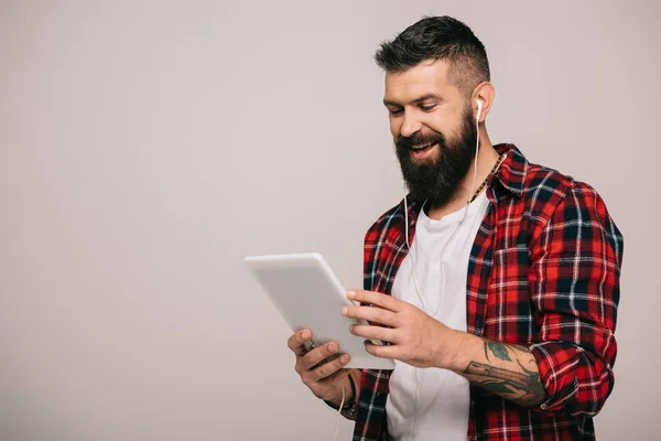 Homme barbu souriant en chemise à carreaux avec écouteurs utilisant une tablette numérique, isolé sur gris — Photo de stock