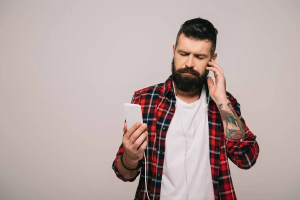 Бородатый мужчина в клетчатой рубашке слушает музыку с наушниками и смартфоном, изолированный на сером — стоковое фото