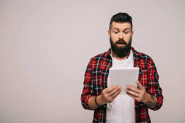 Homme choqué en chemise à carreaux avec écouteurs utilisant une tablette numérique, isolé sur gris — Photo de stock